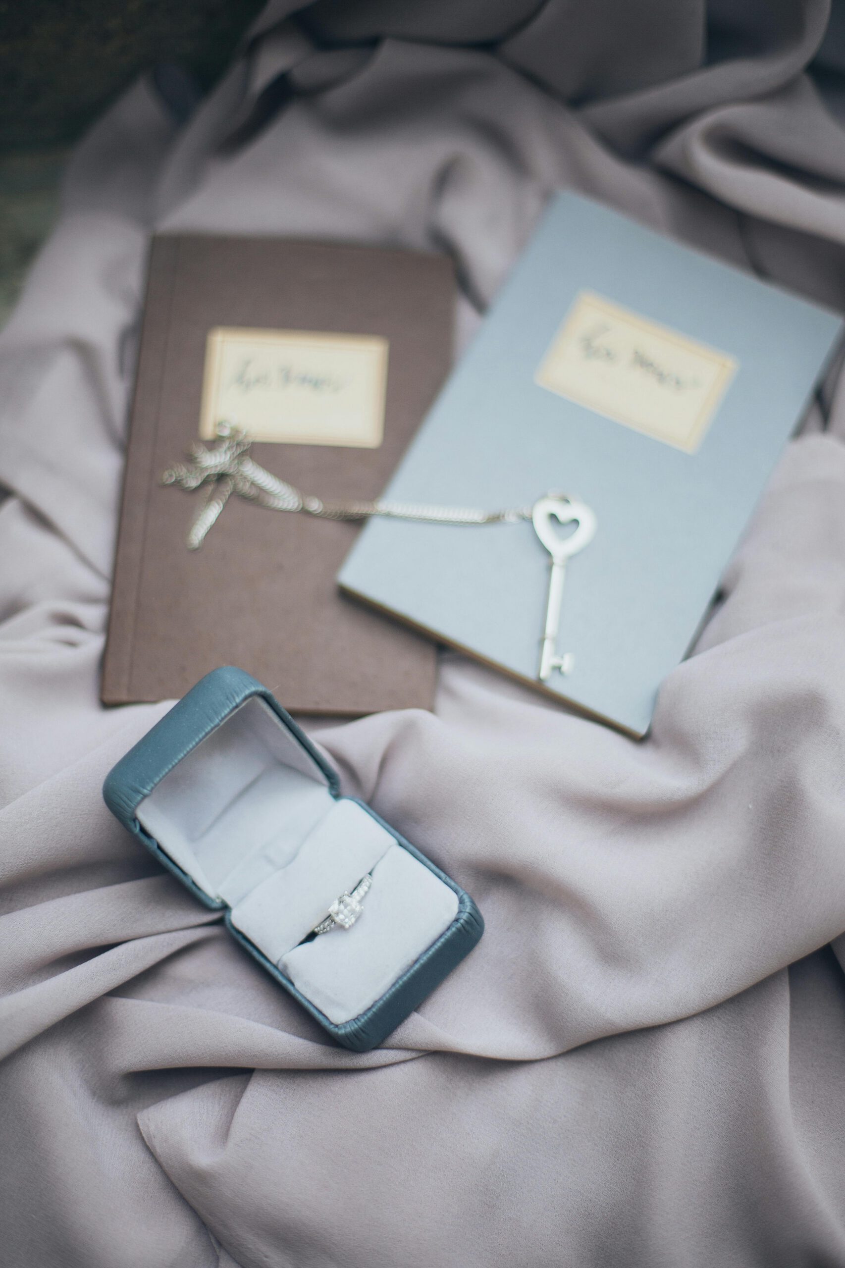 Delicate engagement ring in box beside vintage key and journals on soft fabric backdrop.