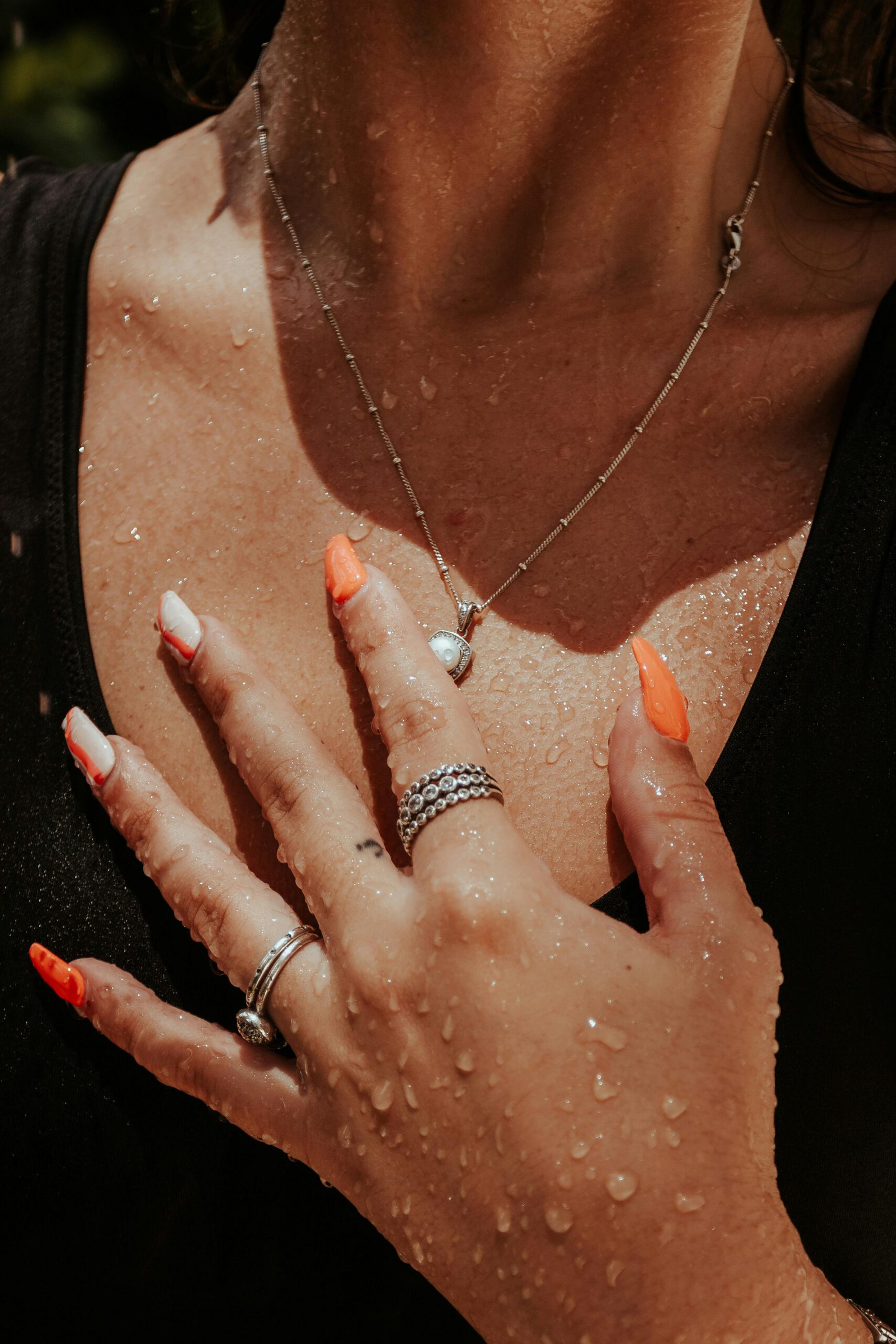 Close-up of a woman's hand with manicured nails, silver rings, and necklace, adorned with water droplets.
